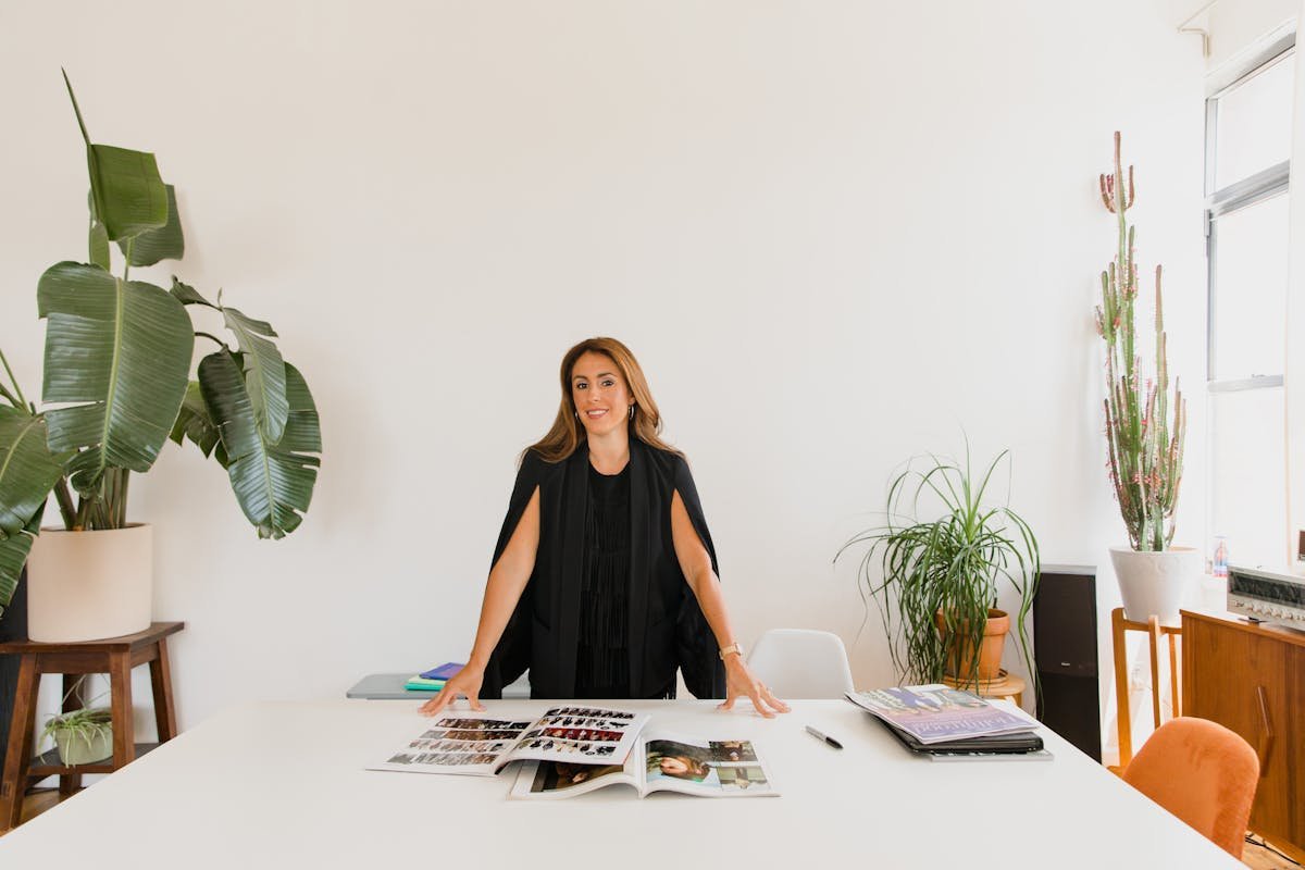 Controle de estoque para pequenos negócios em casa Elegant businesswoman standing confidently at a desk with magazines and indoor plants.