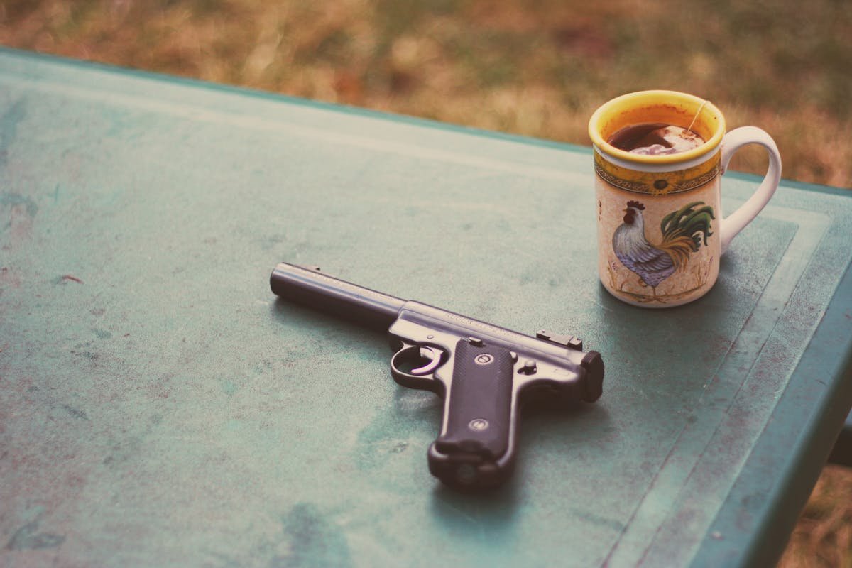 Armas em casa: segurança ou preocupação? A handgun and rooster-themed mug with tea on a green outdoor table.