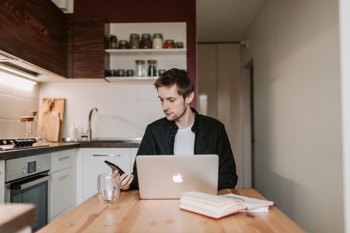 Gerenciando redes sociais em segurança no home office Young male remote employee in casual wear chatting on cellphone while sitting at wooden table in front of open netbook and copybook with glass of water in flat