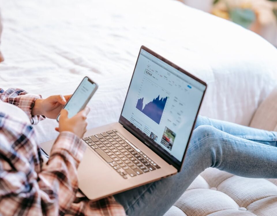 Investimentos em renda fixa: Proteja o futuro da sua casa A woman using a smartphone and laptop to analyze stock market data from home.