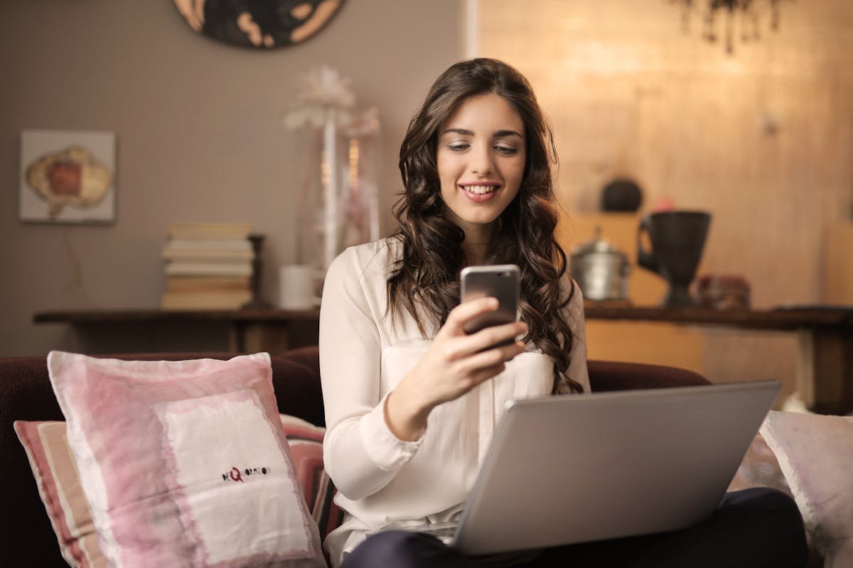 Como sites ajudam na organização de um home office seguro A woman enjoying leisure time using her smartphone and laptop in a cozy living room.
