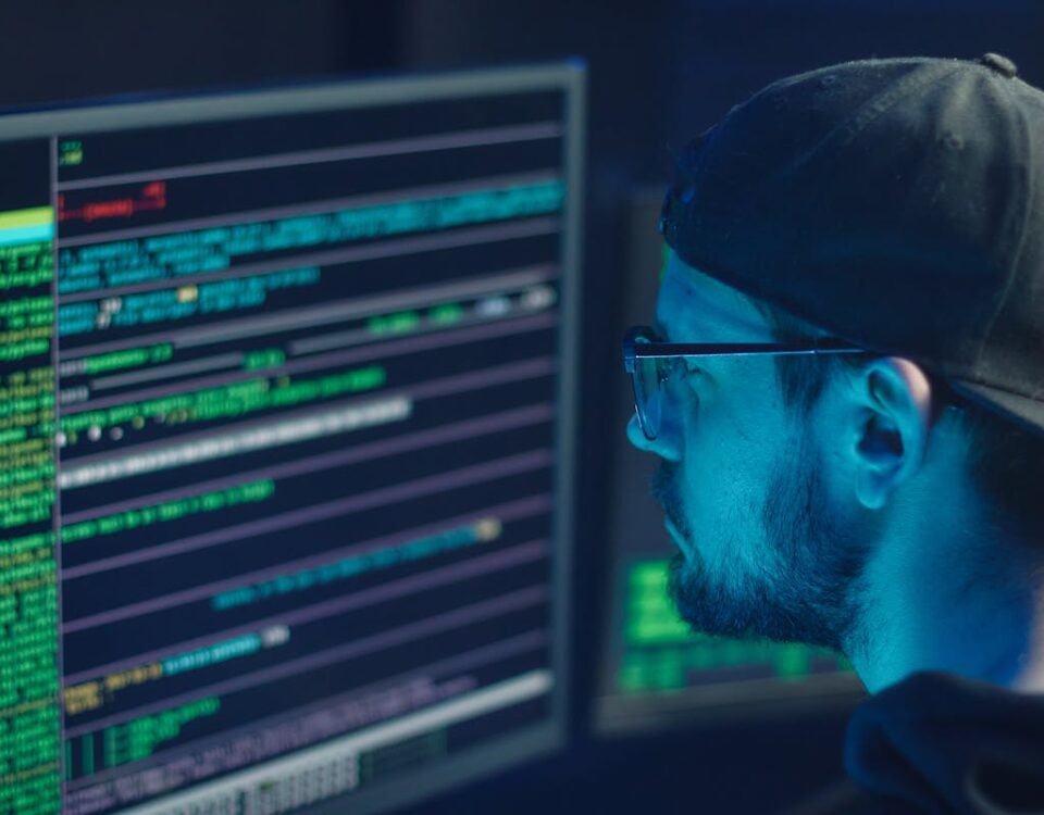Segurança de dados empresariais começa no escritório A cybersecurity expert inspecting lines of code on multiple monitors in a dimly lit office.