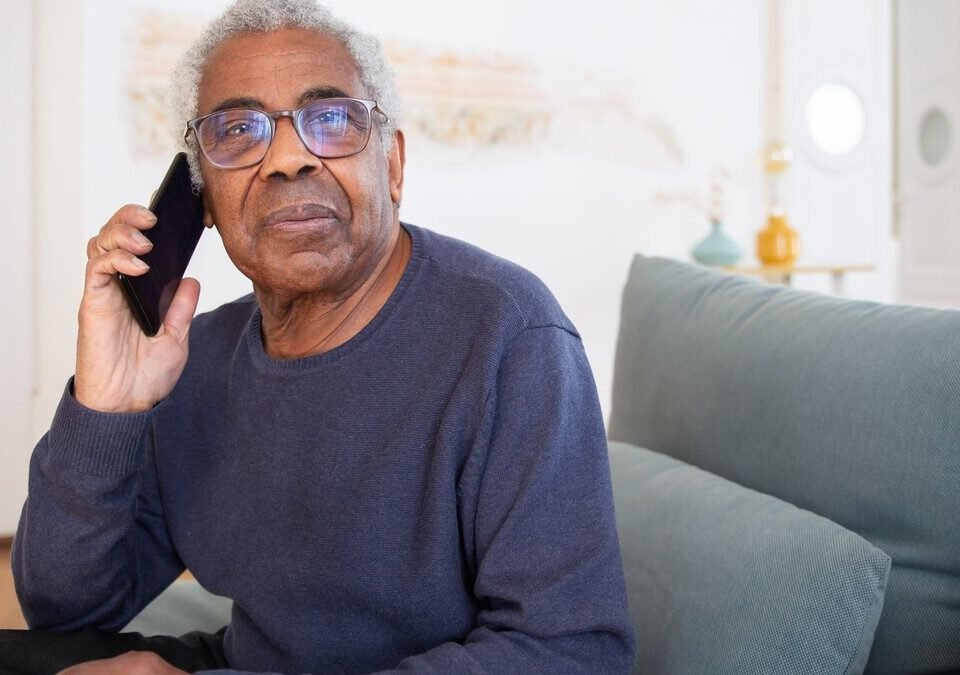 Homem idoso de óculos falando ao celular sentado no sofá de casa