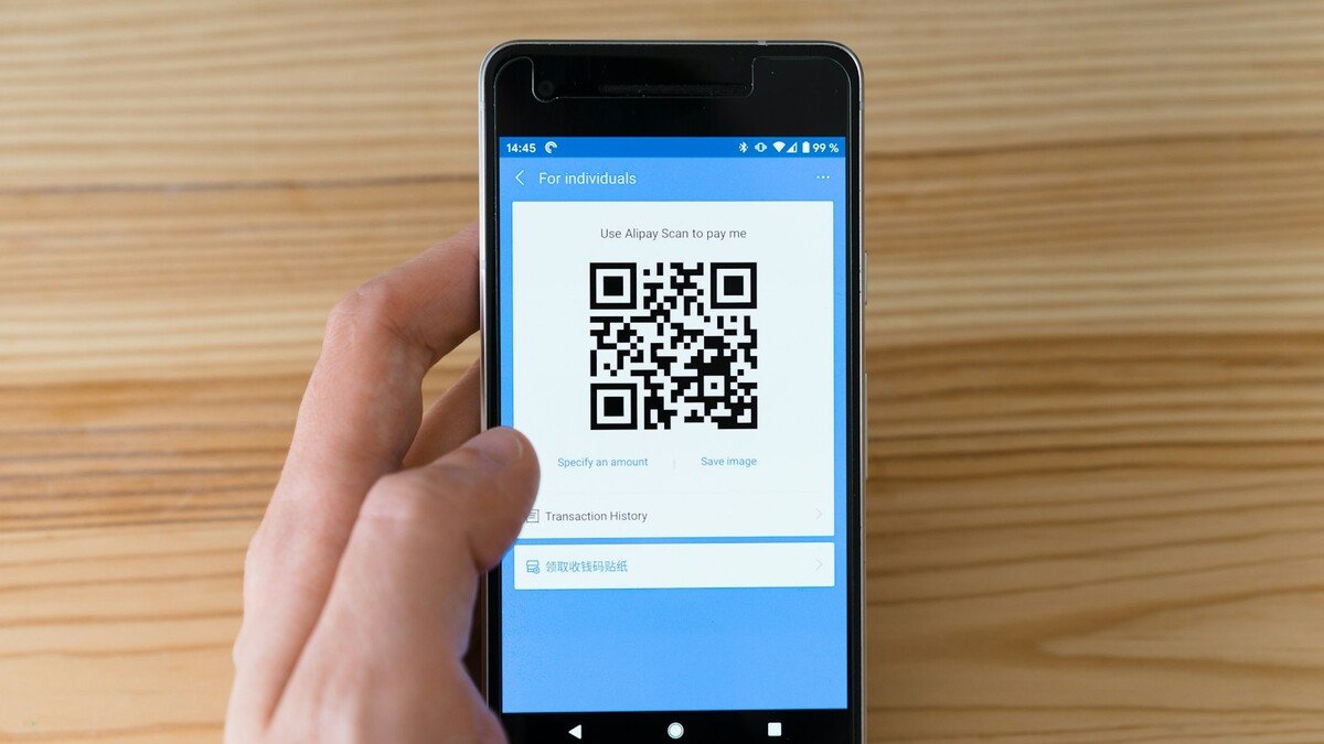 Mão segurando celular Android com QR Code na tela sobre mesa de madeira