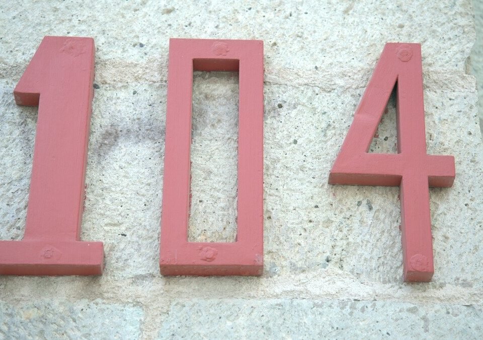 Número 104 em metal vermelho fixado em parede de pedra clara