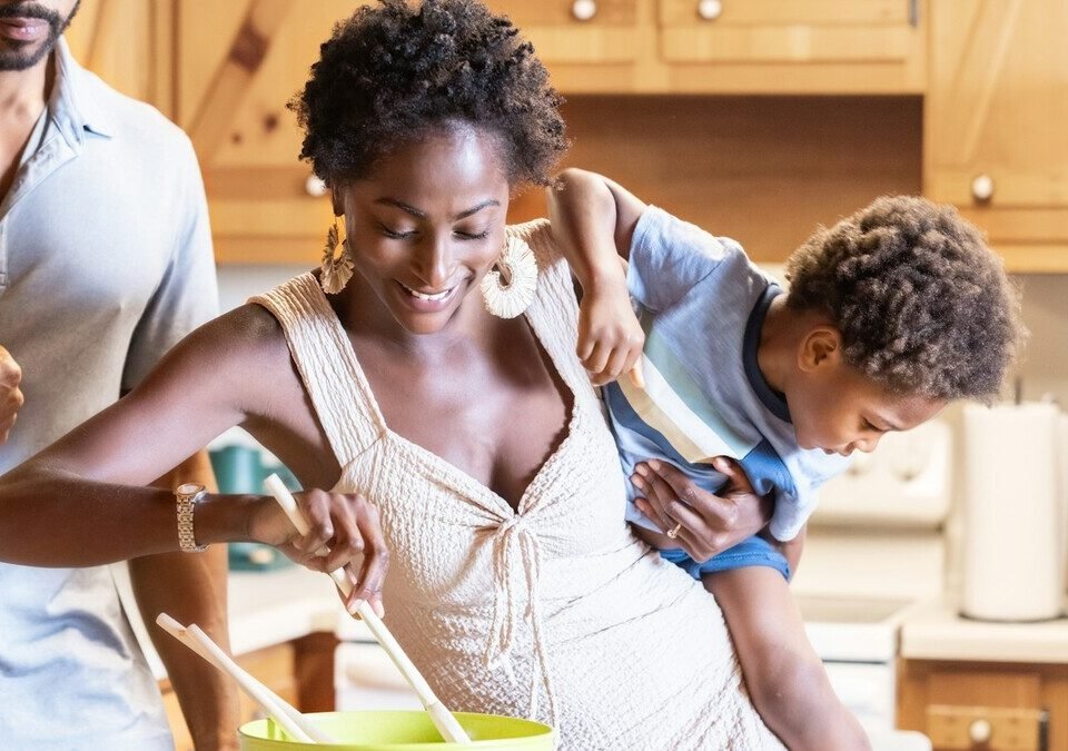 Mulher sorrindo enquanto cozinha, com uma criança nas costas
